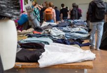 Kleidertauschpartys: Gib Stoff für die Umwelt Kleidung liegt auf Tischen verteilt. Viele Menschen stehen um die Tische. Sie befinden sich auf einer Kleidertauschparty.