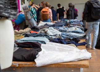 Kleidertauschpartys: Gib Stoff für die Umwelt Kleidung liegt auf Tischen verteilt. Viele Menschen stehen um die Tische. Sie befinden sich auf einer Kleidertauschparty.
