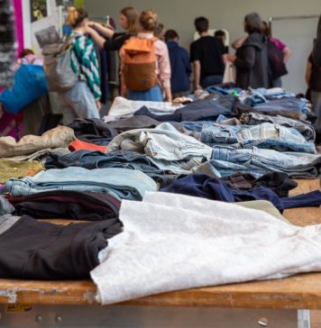 Kleidertauschpartys: Gib Stoff für die Umwelt Kleidung liegt auf Tischen verteilt. Viele Menschen stehen um die Tische. Sie befinden sich auf einer Kleidertauschparty.