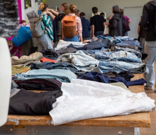 Kleidertauschpartys: Gib Stoff für die Umwelt Kleidung liegt auf Tischen verteilt. Viele Menschen stehen um die Tische. Sie befinden sich auf einer Kleidertauschparty.