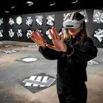Eine Person steht mit ausgestreckten Armen in einem weiten, grauen Raum. Sie trägt eine weiße Virtual-Reality-Brille.