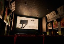 42. Hamburger Kurzfilmfestival Das Bild zeigt einen abgedunkelten Kinosaal. Die Leinwand ist hell beleuchtet. Auf einigen roten Kinosesseln sitzen Personen. Sie sind nur von hinten zu sehen.