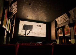 42. Hamburger Kurzfilmfestival Das Bild zeigt einen abgedunkelten Kinosaal. Die Leinwand ist hell beleuchtet. Auf einigen roten Kinosesseln sitzen Personen. Sie sind nur von hinten zu sehen.