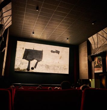 42. Hamburger Kurzfilmfestival Das Bild zeigt einen abgedunkelten Kinosaal. Die Leinwand ist hell beleuchtet. Auf einigen roten Kinosesseln sitzen Personen. Sie sind nur von hinten zu sehen.