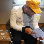 Ein Mann mit einer gelben Cap, Sonnenbrille und weißem Fußball-Deutschland-Trikot sitzt auf einer Bank und hält einen Zettel. Daneben liegen Stapel aus Panini-Sammelstickern.