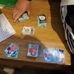 Auf einer Bank liegen 6 Stapel aus Panini-Sammelstickern mit Fußballern. Links liegt ein weißes Blatt auf dem eine Auflistung von Buchstaben steht, die zum Teil abgehakt sind. Eine Hand greift nach einem der Stapel.