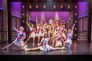 Auf einer rosa beleuchteten Bühne steht das Ensemble von „Natürlich Blond" in einer Endpose nach einer Choreografie. Alle sind wie Cheerleader angezogen und tragen pinkte Schärpen mit der Aufschrift „Team Elle".