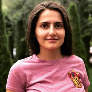 Eine Frau mit braunen, schulterlangen Haaren im Porträt. Sie guckt lächelnd in die Kamera, trägt ein rosa Shirt mit einem Wappen auf der rechten Brust. Im Hintergrund sind Bäume zu erkennen.