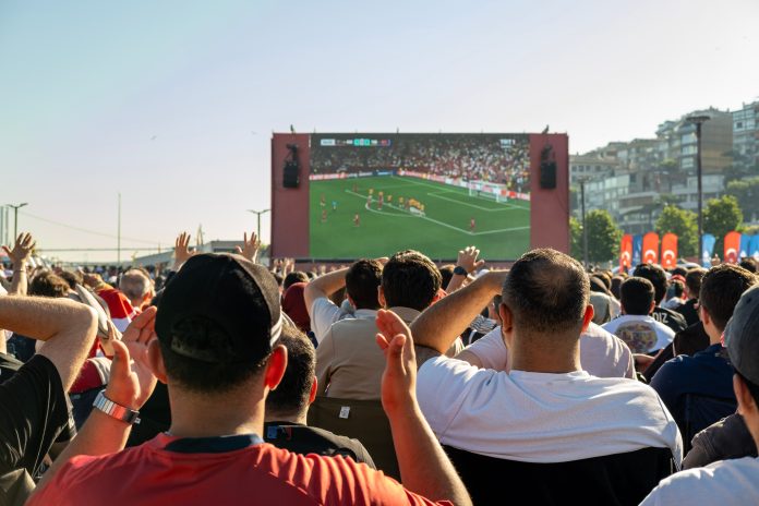 Eine Menge an Menschen steht vor einem großen Bildschirm, auf dem ein Fußball-Spiel gezeigt wird.