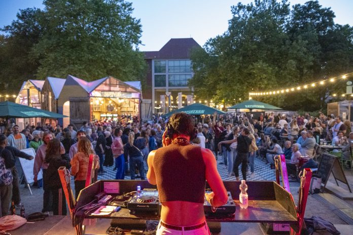 Ein DJ steht auf einer Bühne, mit dem Rücken zur Kamera. Im Hintergrund sieht man Lichterketten und tanzende Menschen. Auf der Altonale 2025.