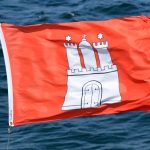 Wo bleibt der Sommer? Rote Hamburg-Flagge weht im Wind. Im Hintergrund ist welliges Wasser zu sehen.