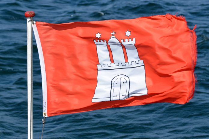 Rote Hamburg-Flagge weht im Wind. Im Hintergrund ist welliges Wasser zu sehen.