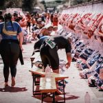 Ironman: Straßensperrungen ab Donnerstag Mehrere Biergarniturbänke stehen auf einer Straße. Ein Mann beugt sich über eine Bank, während links eine Frau in Sportkleidung von der Kamera weggeht. Rechts im Bild hängen Beutel. Im Hintergrund ziehen sich Teilnehmende eines Triathlons um und bereiten sich auf den Wettkampf vor.