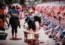 Ironman: Straßensperrungen ab Donnerstag Mehrere Biergarniturbänke stehen auf einer Straße. Ein Mann beugt sich über eine Bank, während links eine Frau in Sportkleidung von der Kamera weggeht. Rechts im Bild hängen Beutel. Im Hintergrund ziehen sich Teilnehmende eines Triathlons um und bereiten sich auf den Wettkampf vor.