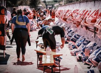 Ironman: Straßensperrungen ab Donnerstag Mehrere Biergarniturbänke stehen auf einer Straße. Ein Mann beugt sich über eine Bank, während links eine Frau in Sportkleidung von der Kamera weggeht. Rechts im Bild hängen Beutel. Im Hintergrund ziehen sich Teilnehmende eines Triathlons um und bereiten sich auf den Wettkampf vor.