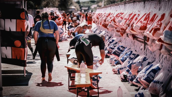 Mehrere Biergarniturbänke stehen auf einer Straße. Ein Mann beugt sich über eine Bank, während links eine Frau in Sportkleidung von der Kamera weggeht. Rechts im Bild hängen Beutel. Im Hintergrund ziehen sich Teilnehmende eines Triathlons um und bereiten sich auf den Wettkampf vor.