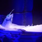 Kampnagel startet Live Art Festival Auf einer Bühne steht eine Badewanne, in der eine Person mit einem Fischkopf-Kostüm sitzt. Die Szene wird von blauem Licht beleuchtet, wodurch eine surreale und geheimnisvolle Atmosphäre entsteht.