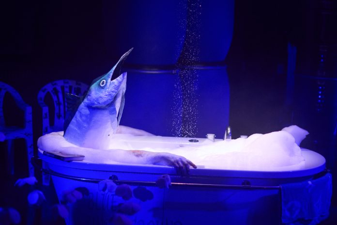 Auf einer Bühne steht eine Badewanne, in der eine Person mit einem Fischkopf-Kostüm sitzt. Die Szene wird von blauem Licht beleuchtet, wodurch eine surreale und geheimnisvolle Atmosphäre entsteht.