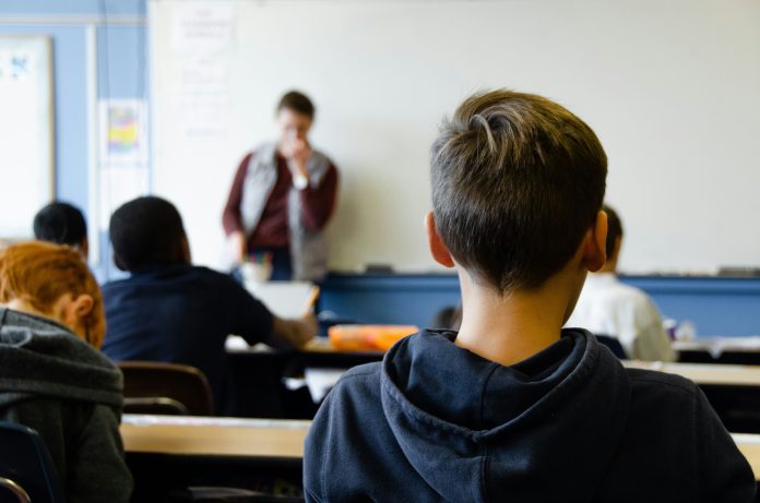 Ein Kind im Vordergrund, welches man von hinten sieht und im Hintergrund ein Lehrer vor einer Tafel. Die GEW möchte, dass Alternativen zum Religionsunterricht ab der ersten Klasse schon angeboten werden.