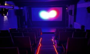 Ein dunkler Kinosaal mit blau-rotem Licht auf der Leinwand. Lichtschläuche umranden den Gang.