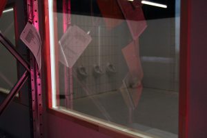 Die nachgebaute Observationskammer der Polizei auf Kampnagel ist mit einer Spiegelscheibe ausgestattet. Sie zeigt einen direkten Einblick in die Toilettenanlage. Vor der Scheibe hängen mehrere Dokumente.
