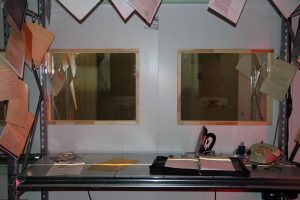 Ein metallischer Bürotisch, auf dem unter anderem Akten und ein Telefon liegen. Weitere Dokumente sind aufgehängt. Der Tisch steht hinter zwei Spiegelscheiben. 