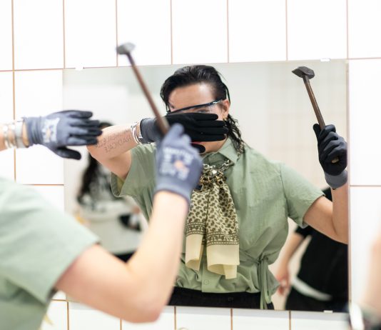 Ein Hammerschlag gegen Toiletten-Überwachung Ausstellungsführerin Sarah Plochl setzt zum Hammerschlag gegen eine Spiegelscheibe in der nachgebauten Toilettenanlage auf Kampnagel an. Sie hält ihre Hand schützend vor ihr Gesicht.