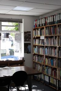 Das Bild zeigt rechts eine Wand voller Bücherregale. Die linke Wand hat Bodentiefe Ladenfenster. Davor steht ein Tisch mit Stühlen.