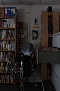 Ein Stapel Stühle steht vor einer Wand an der zwei Plakate hängen. rechts ist ein Aktenschrank und links ein Bücherregal