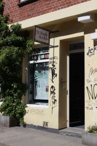 Das Bild zeigt einen Hauseingang. Das Haus ist hellgelb und mit Grafitti bemalt. Die Tür steht offen. Über der Tür hängt ein Schild: St. Pauli-Archiv.