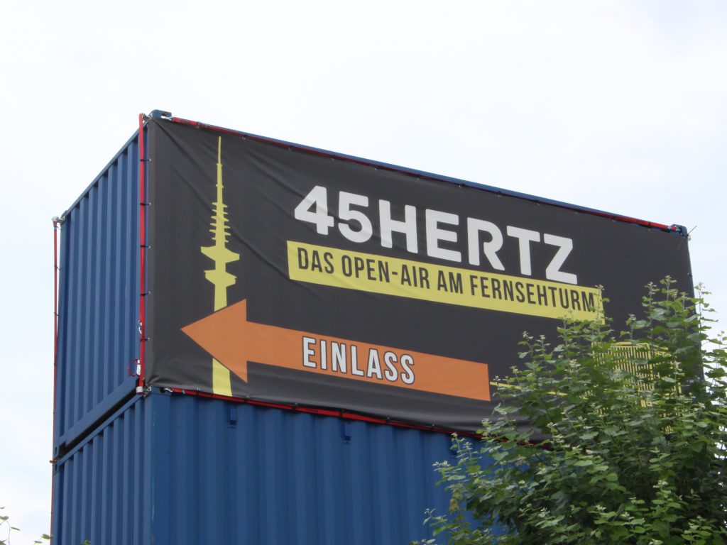 Blaues, rechteckiges Gebäude mit schwarzem Flyer, worauf in weißer Blockschrift steht „45Hertz“. In Gelb ist die Silhoutte eines Fernsehturms abgebildet, darüber in orange ein Pfeil mit der Aufschrift „Eingang“.