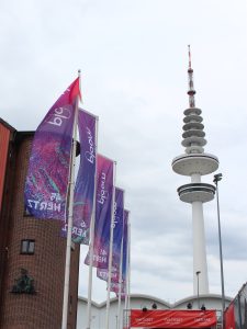 Links sind drei Lila/blaue Flaggen zu erkennen, die hintereinander stehen und im Wind wehen. Rechts im Bild ist der weiße Fernsehturm zu sehen. Unten mittig sieht man einen Eingang. Der Himmel ist hell und grau.