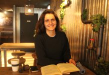 Die albanische Diaspora in Hamburg: Tradition im Wandel Frau in einem Café, vor ihr ein aufgeschlagenes Buch. Sie lächelt in die Kamera. Vor ihr auf dem Tisch sind noch eine Tasse Kaffe und ihr Handy zu sehen.
