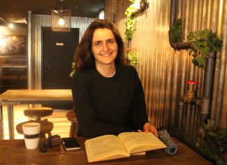 Die albanische Diaspora in Hamburg: Tradition im Wandel Frau in einem Café, vor ihr ein aufgeschlagenes Buch. Sie lächelt in die Kamera. Vor ihr auf dem Tisch sind noch eine Tasse Kaffe und ihr Handy zu sehen.