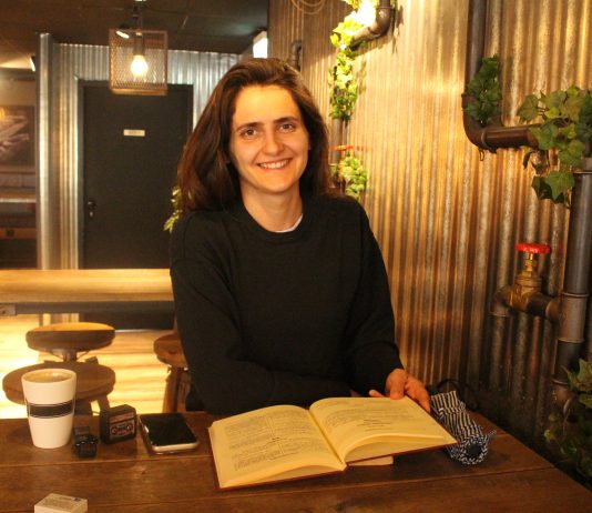 Die albanische Diaspora in Hamburg: Tradition im Wandel Frau in einem Café, vor ihr ein aufgeschlagenes Buch. Sie lächelt in die Kamera. Vor ihr auf dem Tisch sind noch eine Tasse Kaffe und ihr Handy zu sehen.