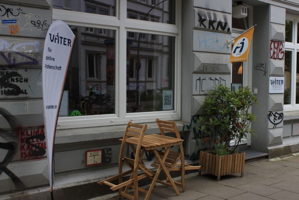 Das Bild zeigt einen ebenerdigen Laden. Vor diesem steht ein Tisch und zwei Stühle auf dem Gehweg. Auf einer Fahne steht: Väterzentrum.