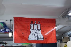 Eine rote Hamburgflagge hängt von der Decke, darauf sehr viele Unterschriften und Zeichnungen.