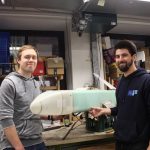 Wettbewerb über den Wolken Vor einer Wand voller Geräte und Schubladenschränke stehen Chris Lucas Siewert und Fabian Fabian Kühn, zwei Mitglieder des Vereins Neues Fliegen, mit einem Modellflugzeug.