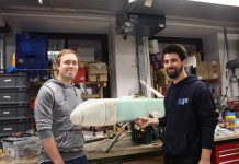 Wettbewerb über den Wolken Vor einer Wand voller Geräte und Schubladenschränke stehen Chris Lucas Siewert und Fabian Fabian Kühn, zwei Mitglieder des Vereins Neues Fliegen, mit einem Modellflugzeug.