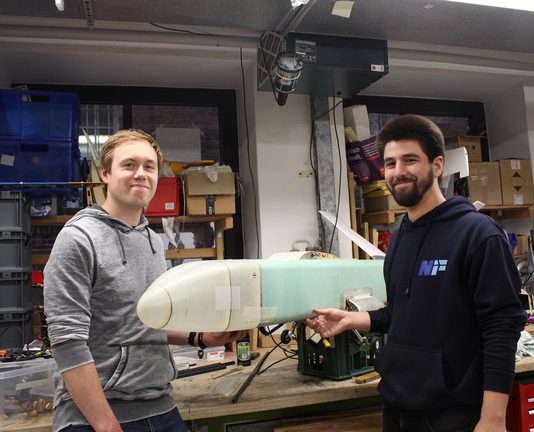 Wettbewerb über den Wolken Vor einer Wand voller Geräte und Schubladenschränke stehen Chris Lucas Siewert und Fabian Fabian Kühn, zwei Mitglieder des Vereins Neues Fliegen, mit einem Modellflugzeug.