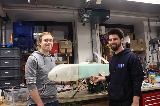 Chris Lucas Siewert und Fabian Kühn vom Verein Neues Fliegen mit einem selbstgebauten Modellflugzeug. Foto: Mia Wietkamp Vor einer Wand voller Geräte und Schubladenschränke stehen Chris Lucas Siewert und Fabian Fabian Kühn, zwei Mitglieder des Vereins Neues Fliegen, mit einem Modellflugzeug.
