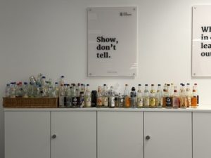 Vor einem „Show don't tell" Plakat im Newsroom stehen sehr viele Pfandflaschen auf einem Weißen Regal.