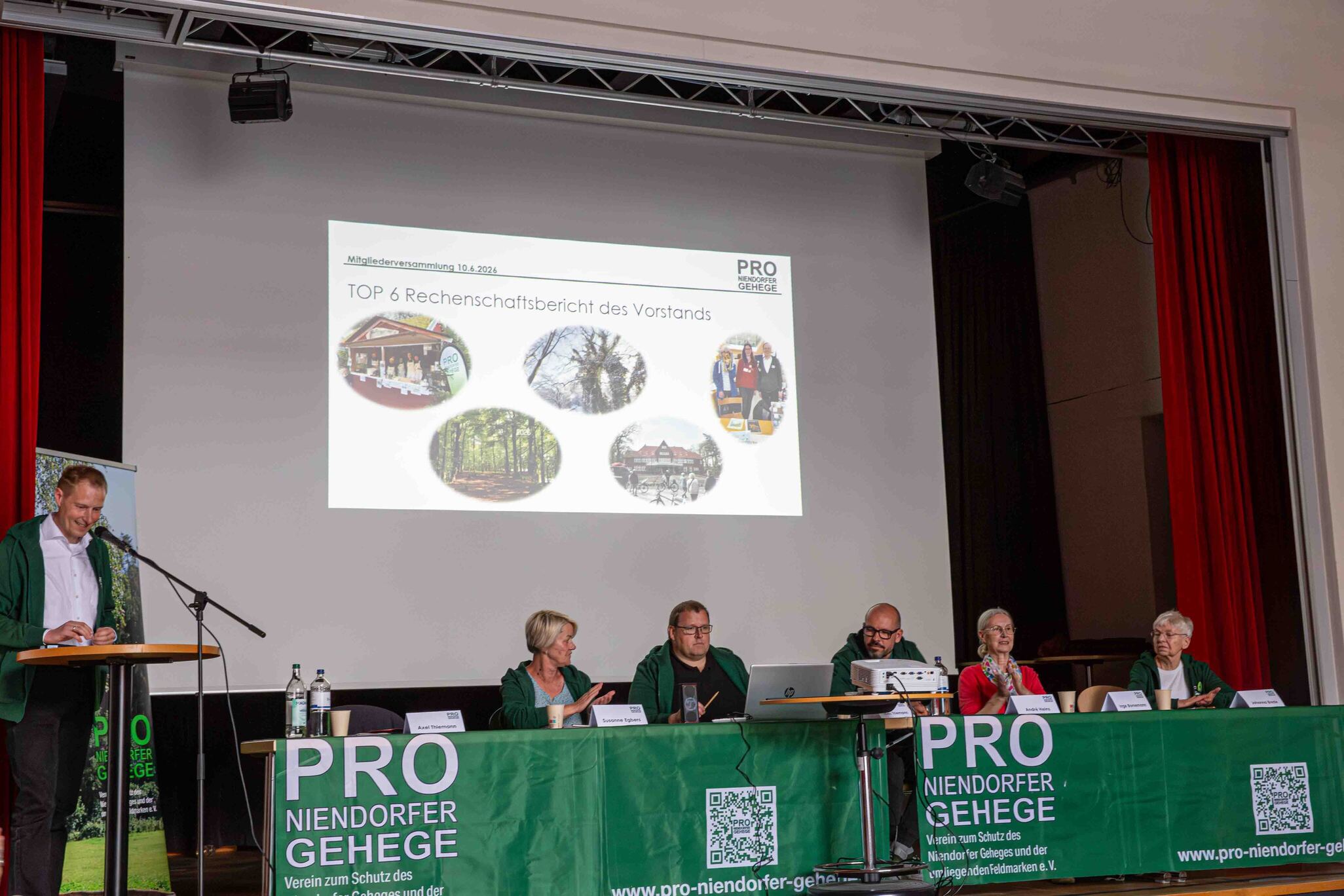 In einer Aula sitzen mehrere ältere Personen vor einer Bühne. Auf der Bühne ist ein langer Tisch, hinter dem die sechs Vorstandsmitglieder des Vereins „Pro Niendofer Gehege" sitzen. Der Tisch hat ein grünes Banner, auf dem „Pro Niendorfer Gehege" steht. Auf der Leinwand hinter dem Vorstand ist die Tagesordnung zu sehen.