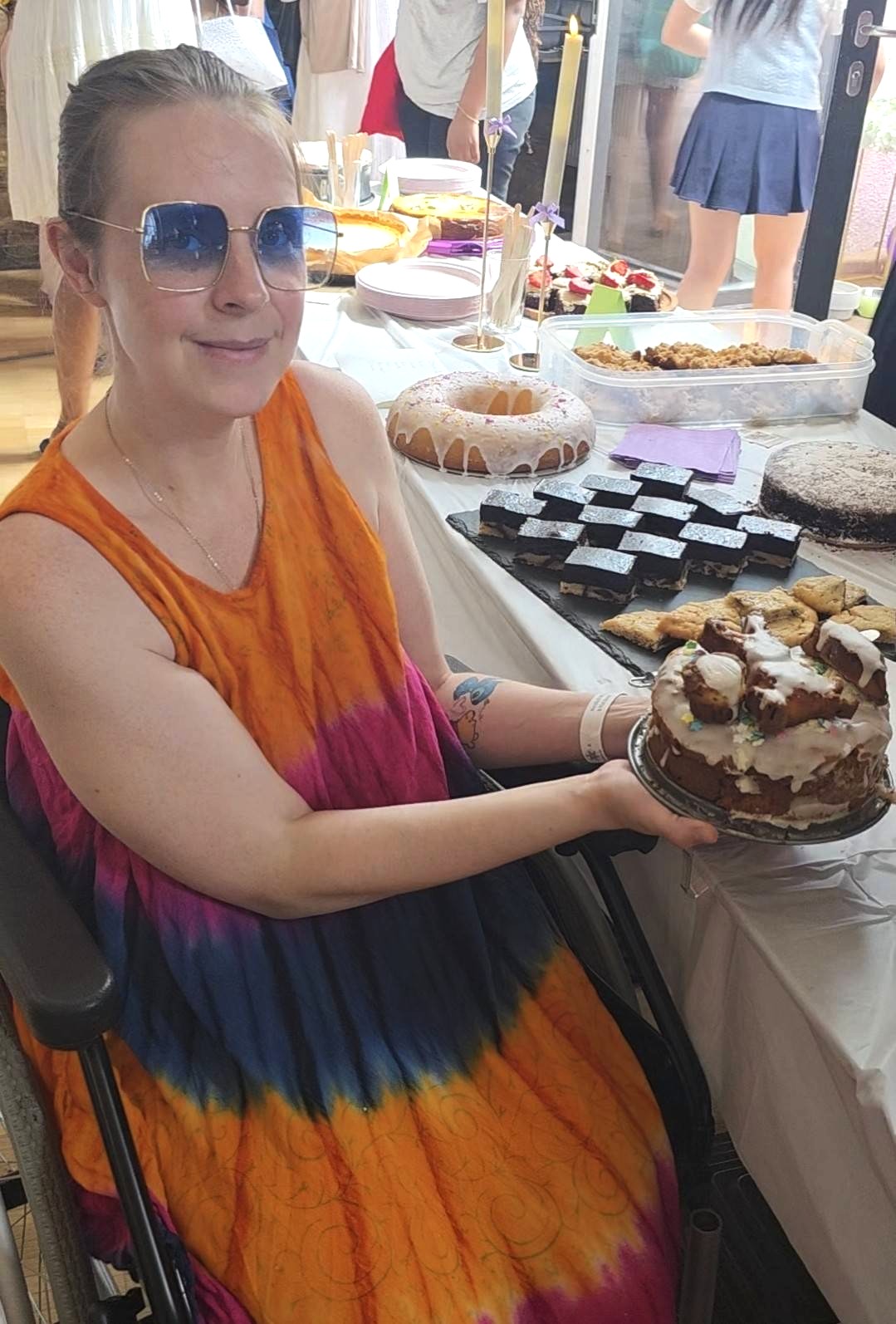 Nina sitzt mit einer Sonnebrille in einem orange-lilanem Kleid in einem Rollstuhl neben einem der Kuchentische. Sie hält eiinen Kuchen in den Händen, der auf einem Teller steht. Der Kuchen hat eine Schicht aus Zuckerglasur.