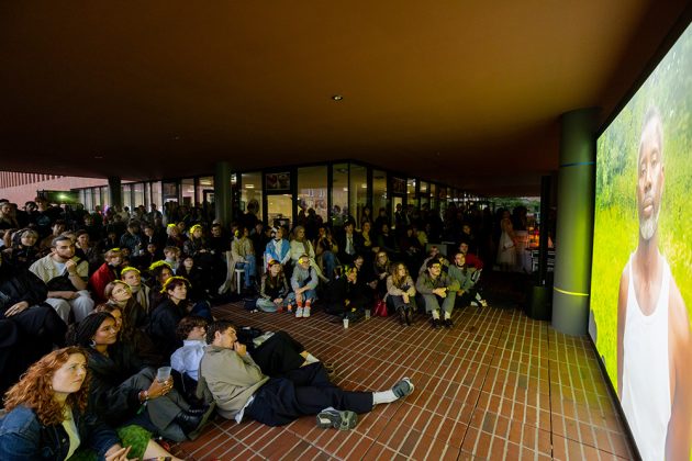 Viele Menschen gucken auf einen Bildschirm. Es ist dunkel, einige Menschen sitzen auf dem Boden.