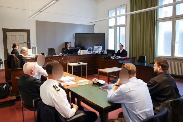 In einem Gerichtsaal sitzen Personen an Tischen zueinander, wobei drei Gesichter zesniert sind.