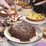 “Kuchen ist für jeden da, oder?” Auf einem Tisch liegen verschiedene Kuchen. In der Mitte liegt ein Schokoladenkuchen, dahinter andere Kuchen mit Creme-Schicht. mehrere Menschen schneiden die Kuchen gleichzeitig an.