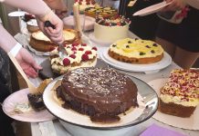 “Kuchen ist für jeden da, oder?” Auf einem Tisch liegen verschiedene Kuchen. In der Mitte liegt ein Schokoladenkuchen, dahinter andere Kuchen mit Creme-Schicht. mehrere Menschen schneiden die Kuchen gleichzeitig an.