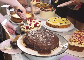 “Kuchen ist für jeden da, oder?” Auf einem Tisch liegen verschiedene Kuchen. In der Mitte liegt ein Schokoladenkuchen, dahinter andere Kuchen mit Creme-Schicht. mehrere Menschen schneiden die Kuchen gleichzeitig an.