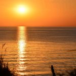 Nord- und Ostsee: Meeresoberfläche hat sich im Juni schnell erwärmt Blick auf die Ostsee. Aufgehende Sonne über dem Horrizont. Schattige Gräser im Vordergrund.